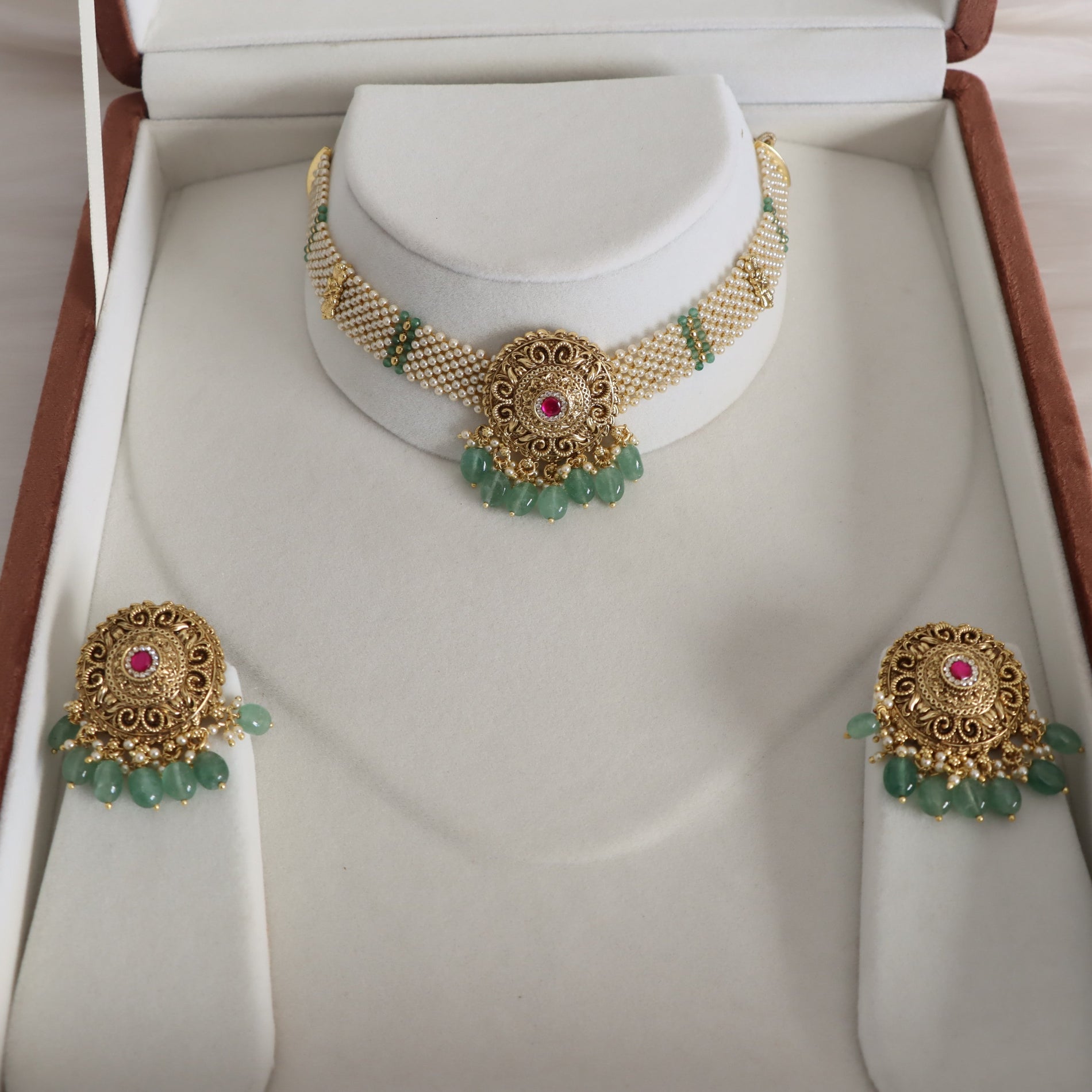Gold-toned traditional jewelry set displayed in an open presentation box, including a choker necklace, matching earrings, and small dangling accents, with green bead drops and red center stones.