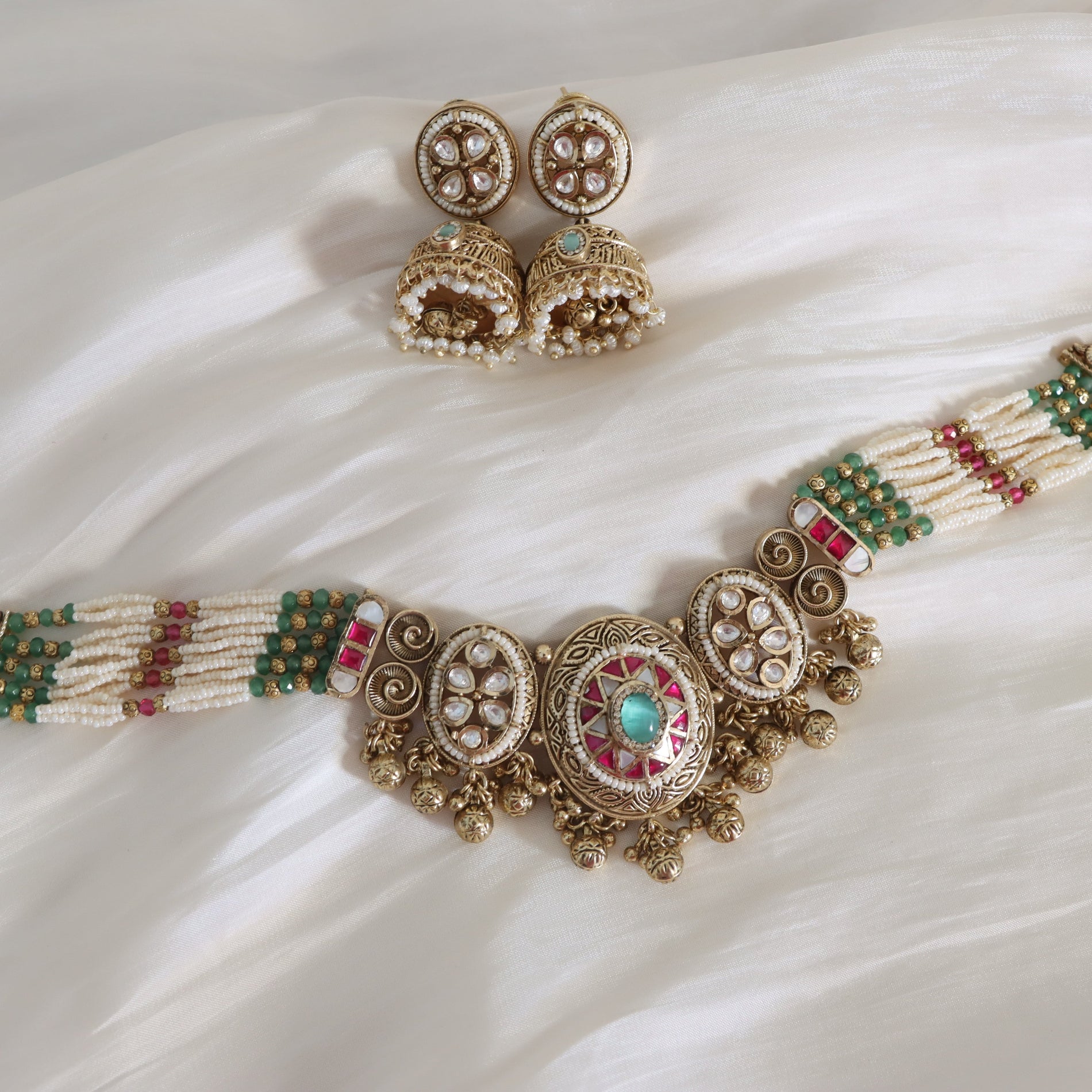 Ornate gold necklace with green and red stones and matching earrings on a white fabric background