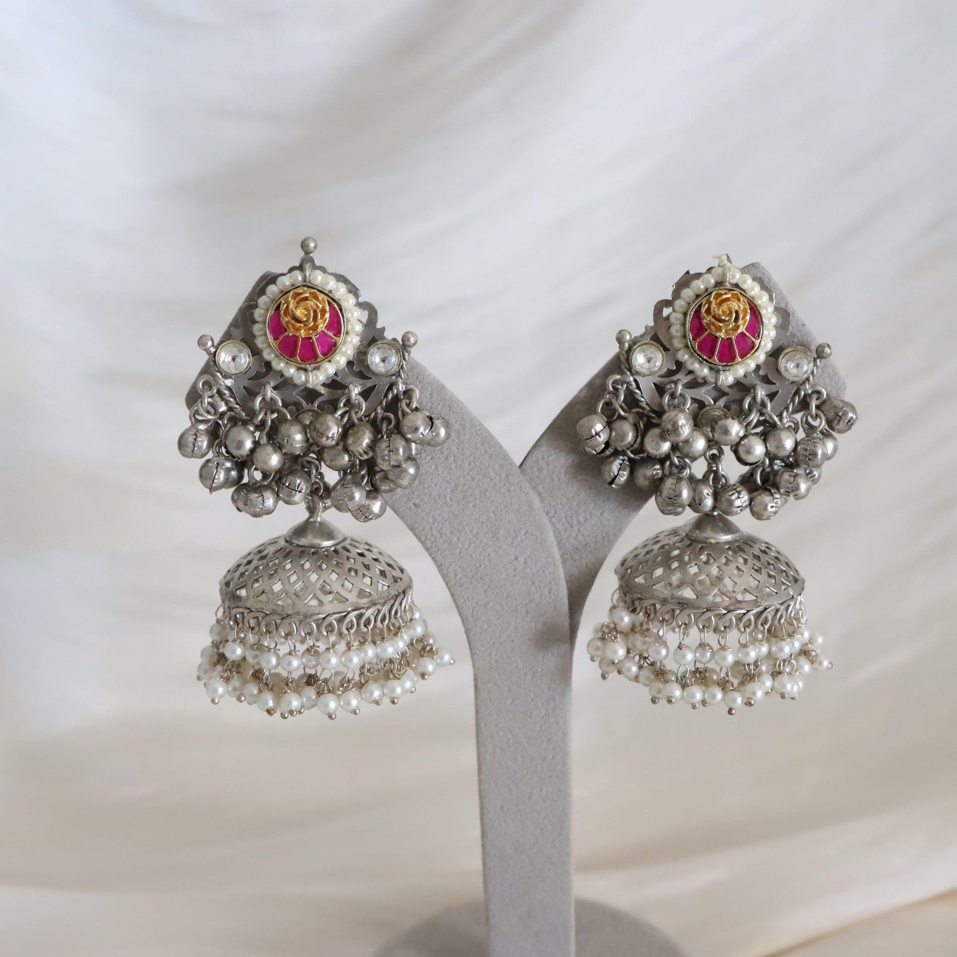 A pair of ornate silver-toned jhumka earrings with pink and yellow floral enamel details and dangling pearl-like beads, displayed on a gray stand against a soft white fabric background.