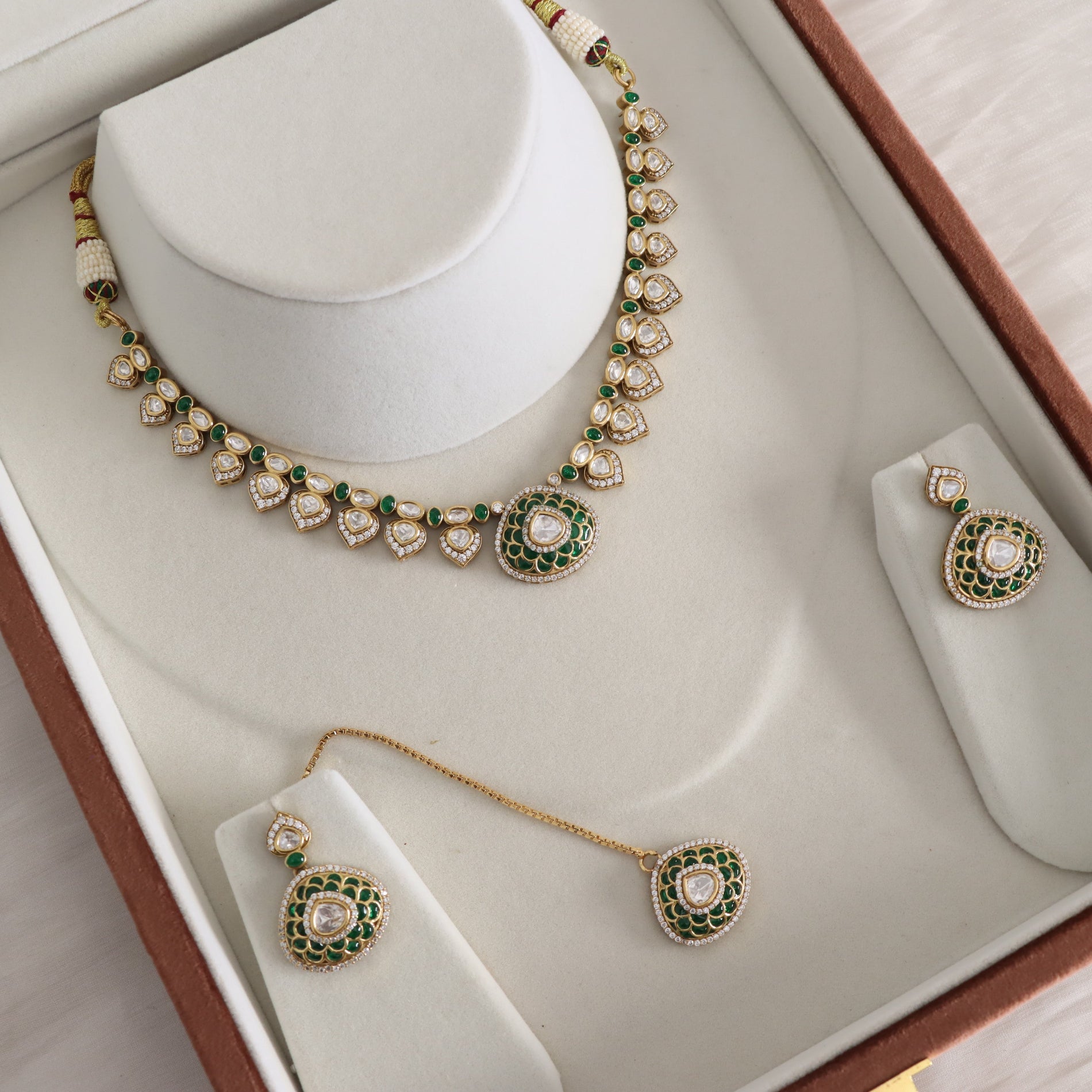 A gold-toned traditional jewelry set displayed in an open presentation box, including a necklace, matching earrings, and a dangling pendant, all decorated with green stones and clear stones.