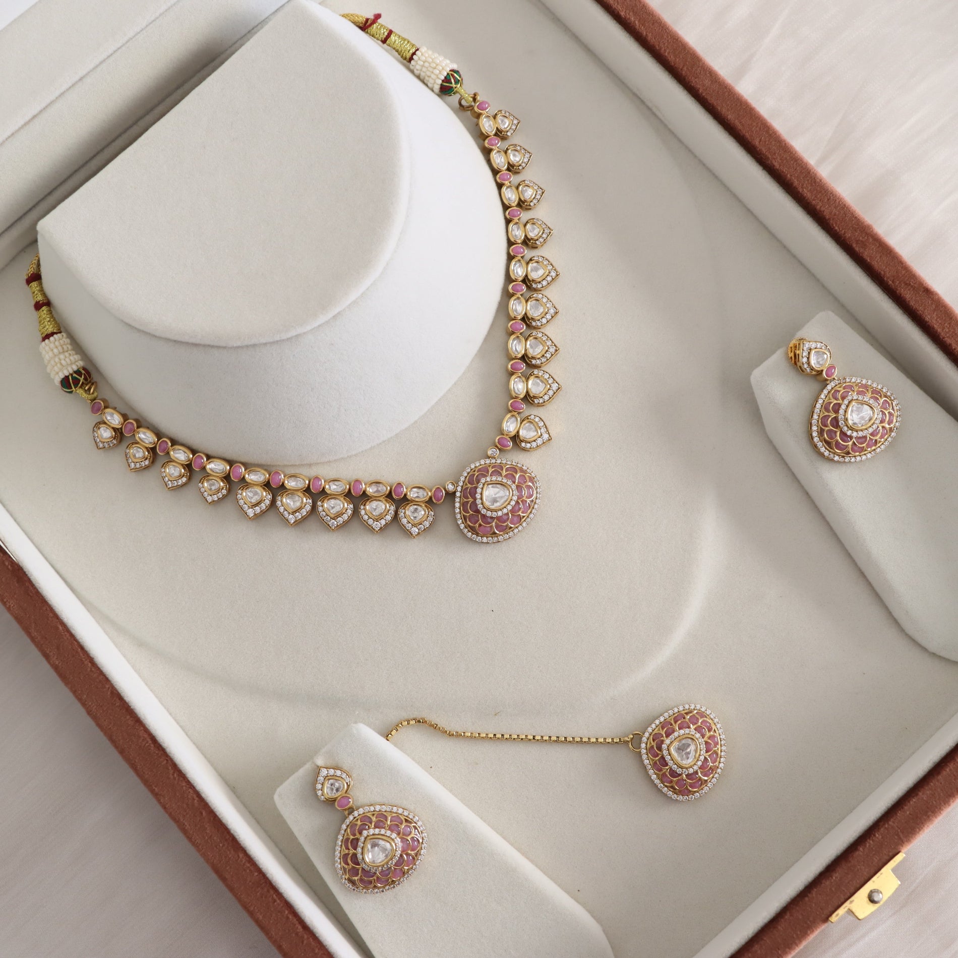 Gold and pink stone jewelry set displayed on a white jewelry box, including a necklace on a white stand, matching earrings on small stands, and a maang tikka with a connecting chain.