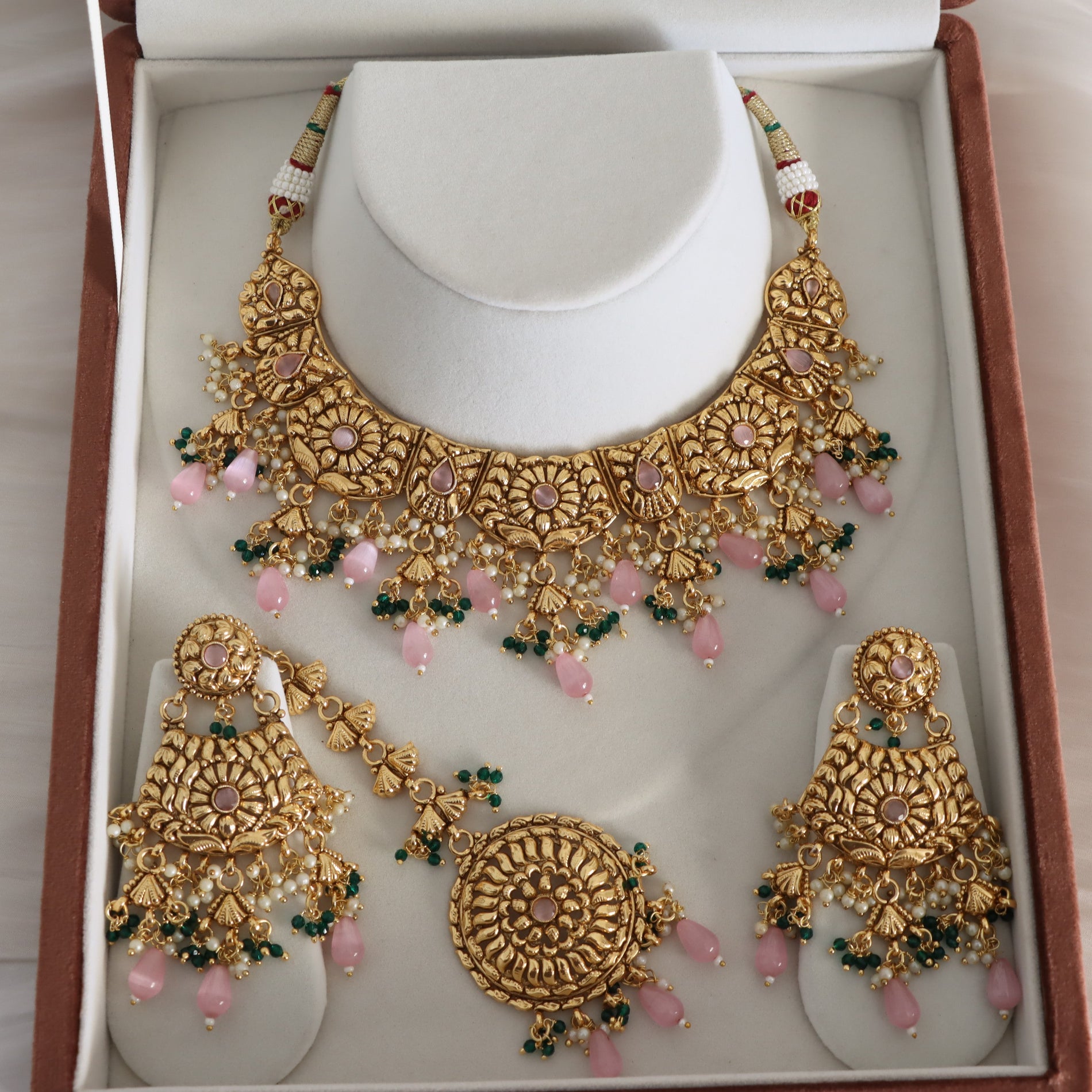 Gold-toned traditional Indian jewelry set displayed in a presentation box, including a choker necklace, earrings, and a matching maang tikka, decorated with pink stones and green bead accents.
