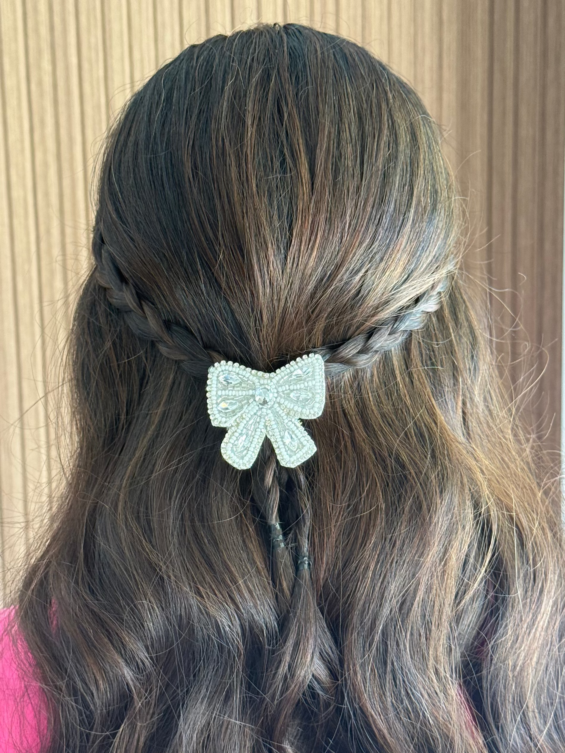 A close-up back view of a person with long dark brown hair styled in a half-up braided ponytail adorned with a light blue rhinestone bow, set against a beige wood-paneled background.