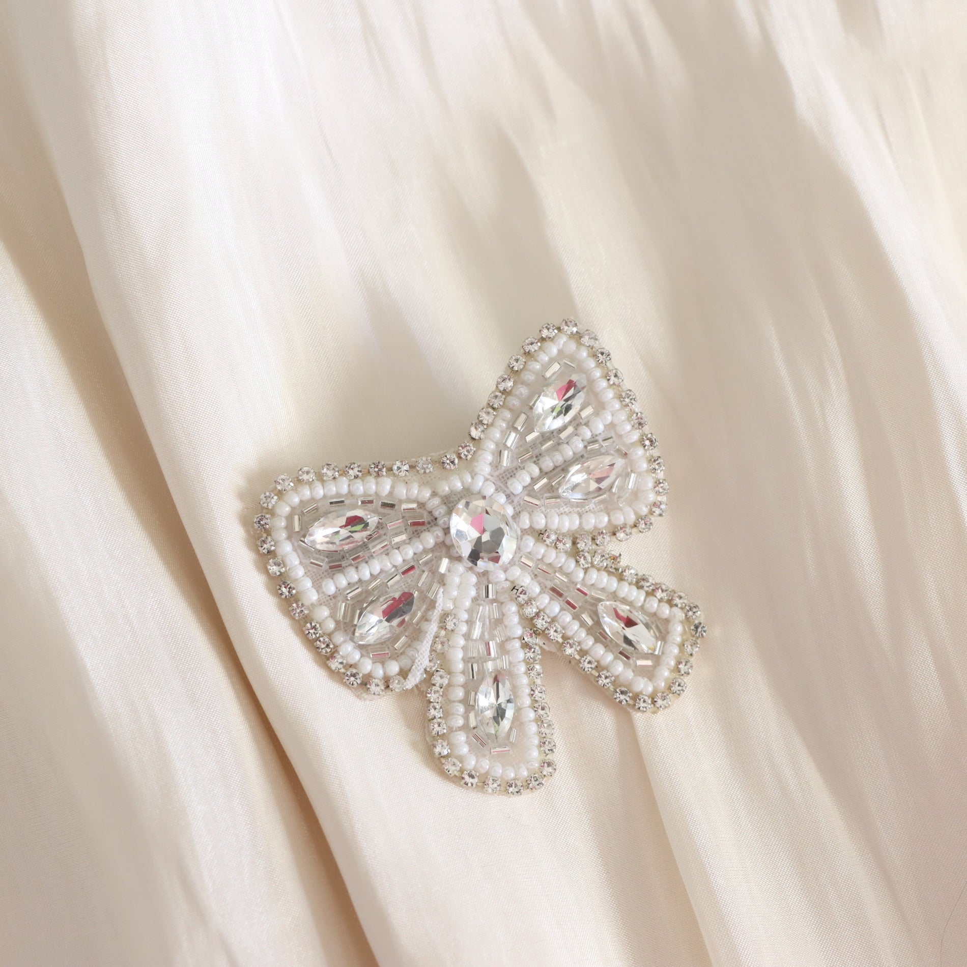 A bow-shaped beaded brooch with clear rhinestones and a central crystal, displayed on a soft cream-colored fabric background.