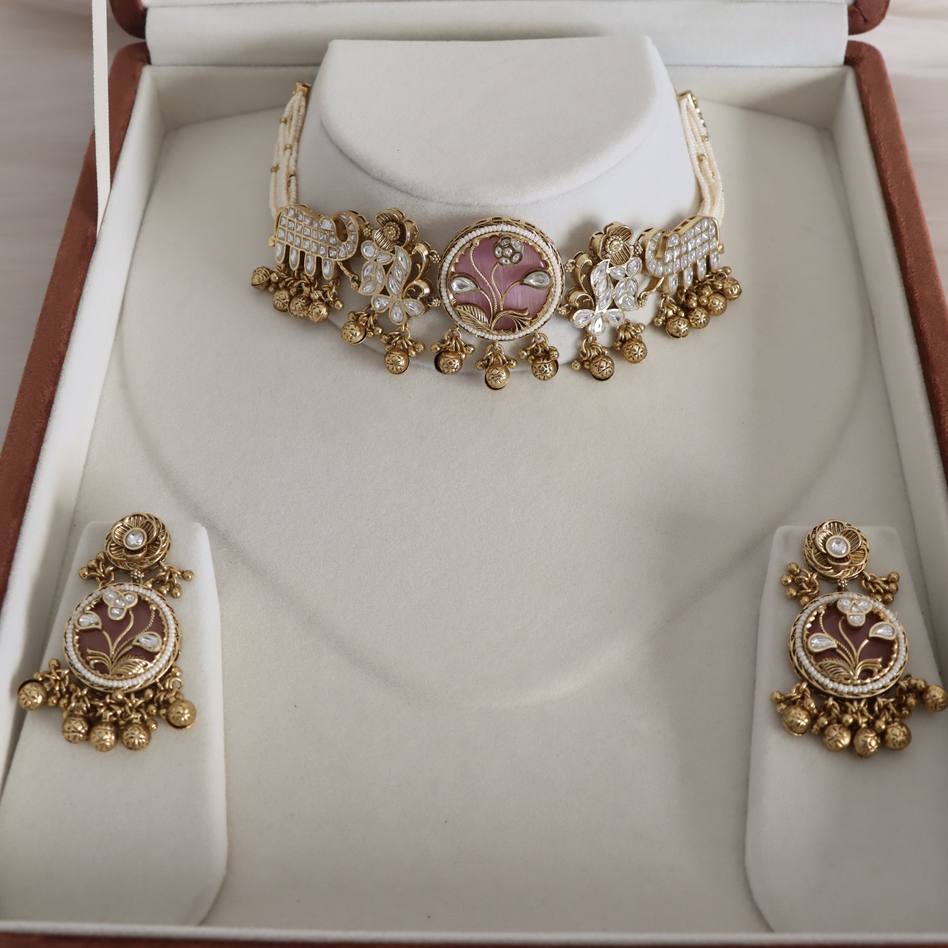 An ornate gold-toned jewelry set displayed inside a cream jewelry box. The set includes a choker necklace and matching drop earrings, both decorated with gold filigree, white faux pearls, and central oval pink enamel medallions.