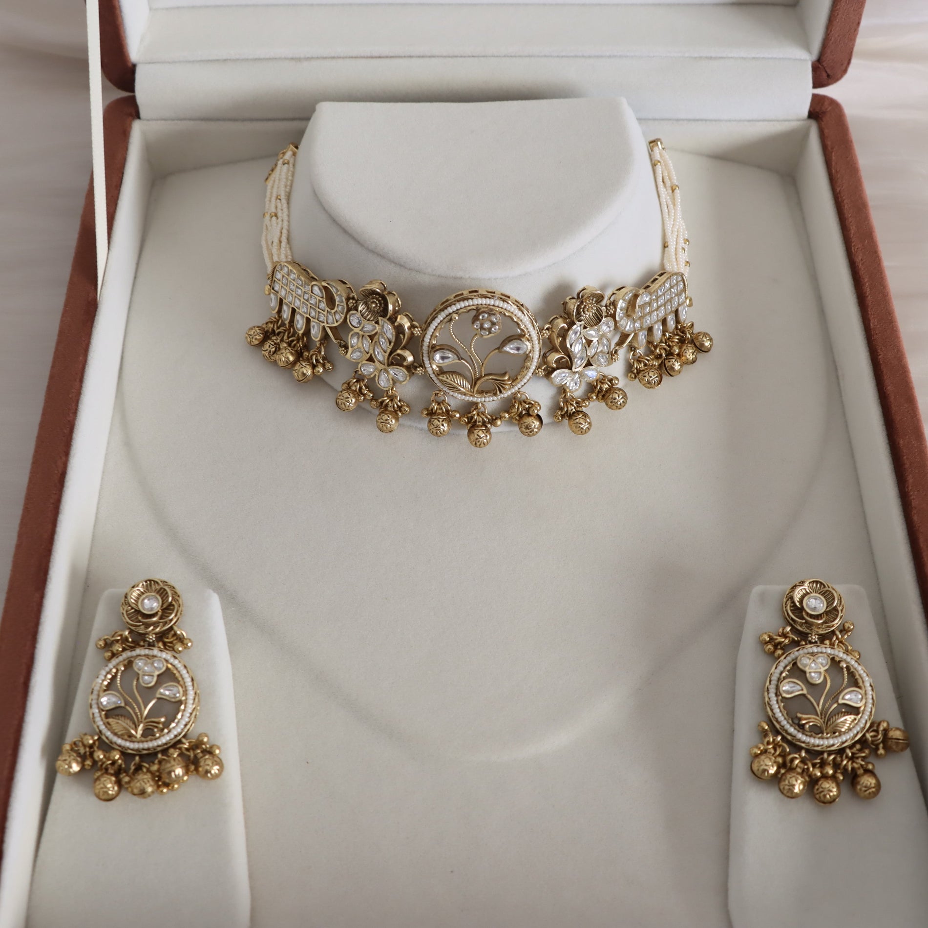 Gold-toned traditional Indian jewelry set including a choker necklace, earrings, and a bracelet, displayed in an open presentation box with cream-colored velvet inserts.