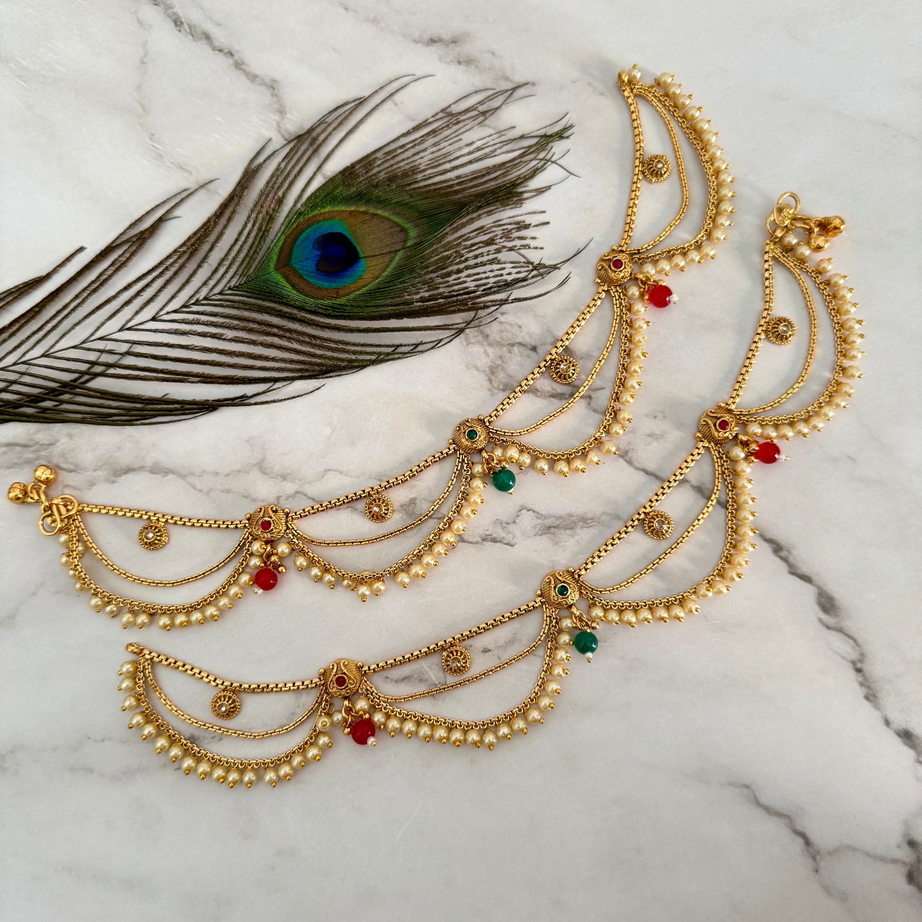 Gold anklet with red and green gemstones on a marble surface with a peacock feather.