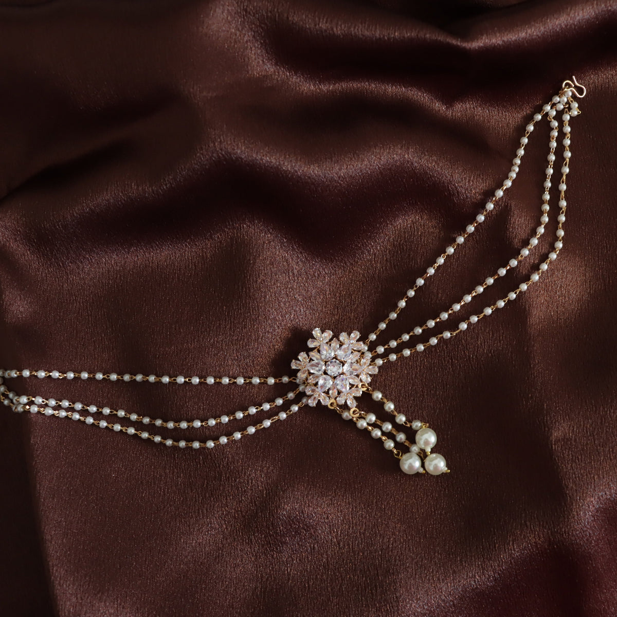 A double-strand pearl choker necklace with a central crystal-embellished floral brooch and dangling pearl accents, displayed on a draped dark brown satin fabric.
