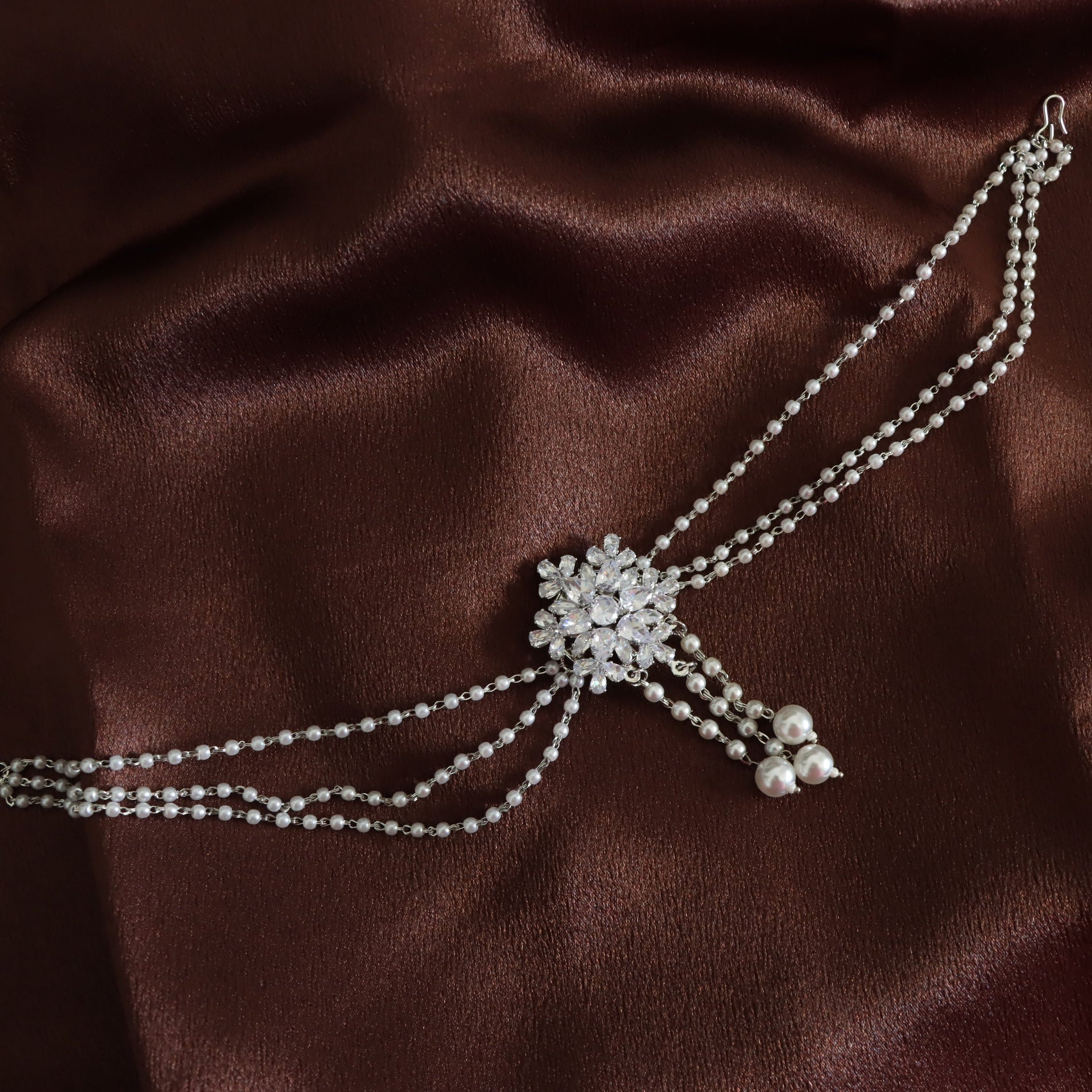 A silver-toned double-strand chain choker with a central crystal floral brooch and dangling pearl beads, displayed on a draped dark brown satin fabric.