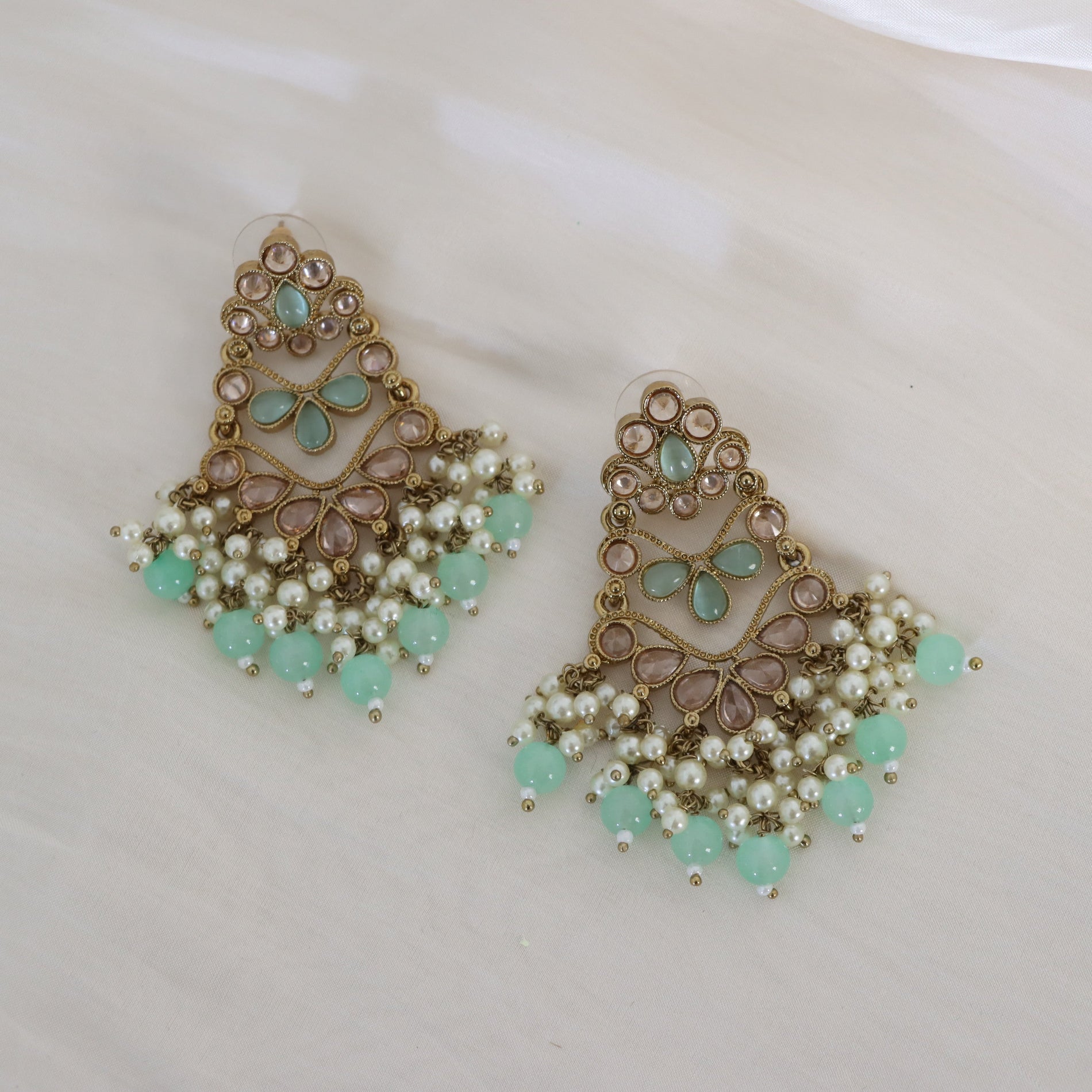 A pair of ornate gold-toned statement earrings with light green stone accents, white faux pearls, and dangling mint green beads, displayed on a light cream surface.