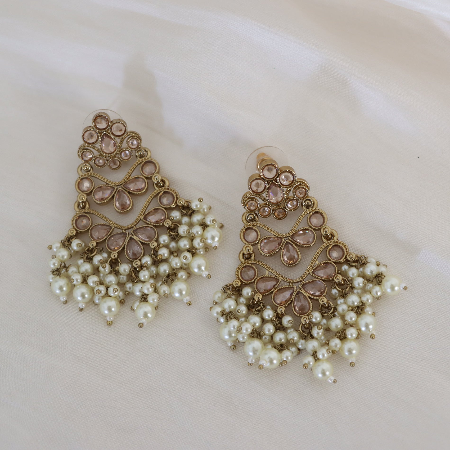 A pair of ornate gold-toned statement earrings with pink teardrop crystals and cascading white faux-pearl fringe, displayed on a soft cream-colored fabric background.