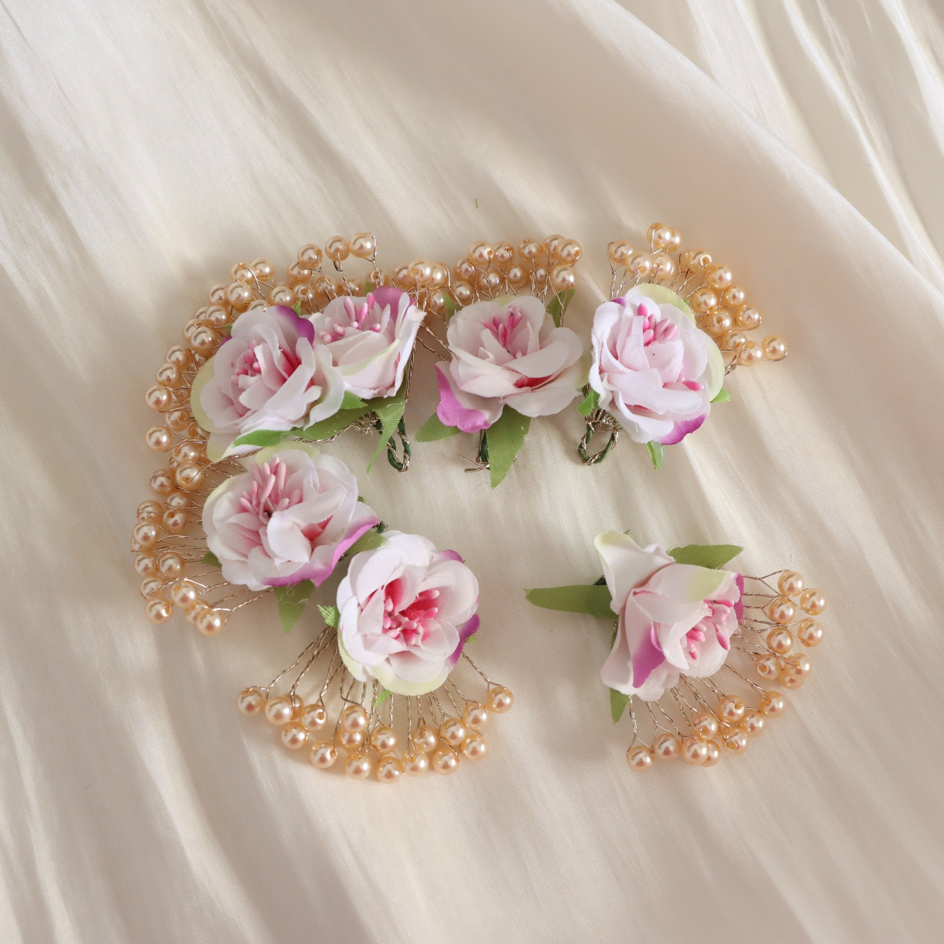 A set of seven floral hair clips arranged on a draped cream fabric background. Each clip features a pink and white artificial rose with green leaves, accented by clusters of small cream pearls.