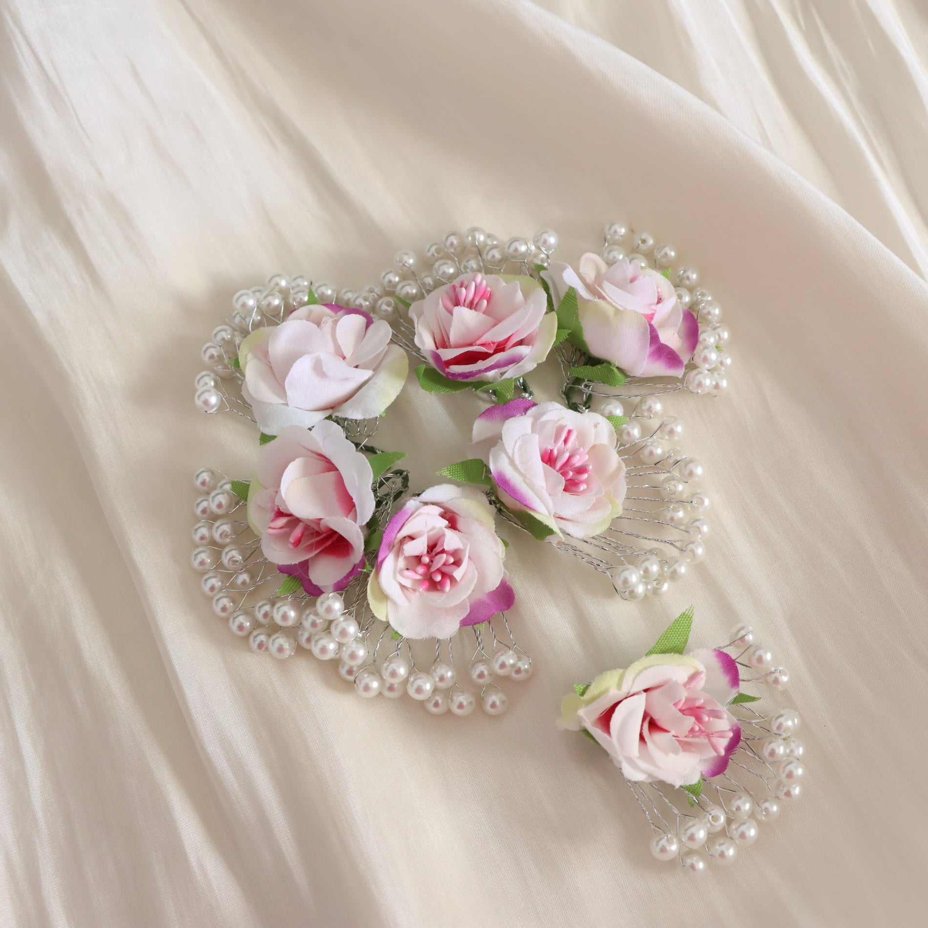 A set of five bridal hair clips decorated with pink and white faux roses, green leaves, and clusters of white faux pearls, arranged on a draped cream satin fabric.