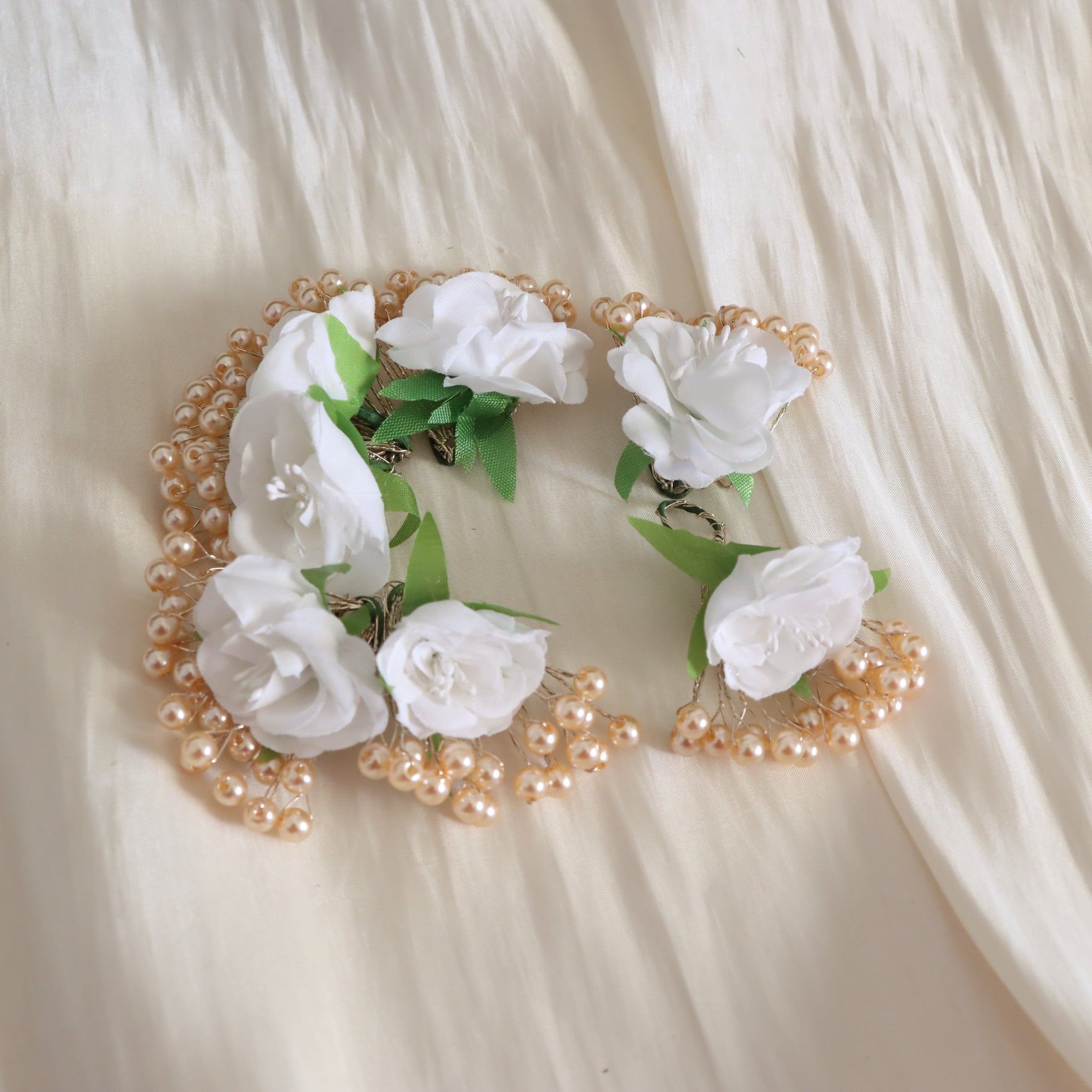 A white floral hair accessory with white fabric roses, green leaves, and strands of small beige faux pearls, laid flat on a cream-colored fabric background.