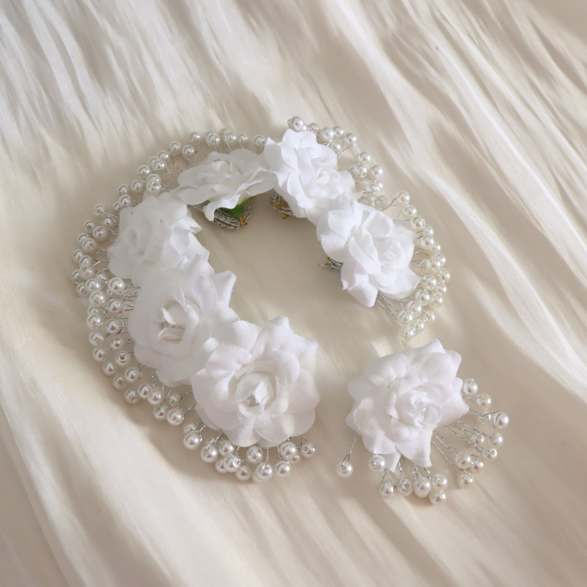 White floral hair accessory with faux white roses and pearl beads, displayed on a cream-colored draped fabric background.