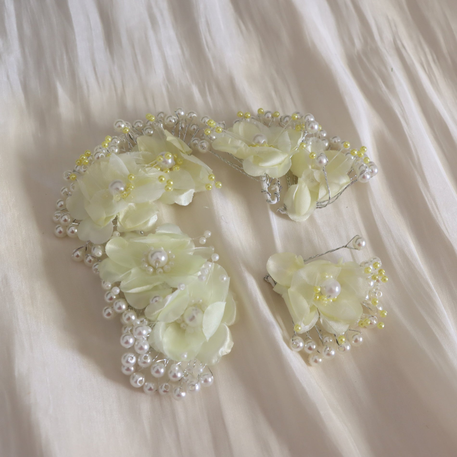 A bridal hair accessory set featuring a headband and matching hair clips, decorated with pale yellow satin flowers, faux pearls, and small yellow beads, displayed on a draped cream satin fabric.